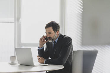 Mature businessman using laptop and cell phone on table in office - MOEF02530