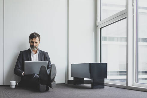 Reifer Geschäftsmann im Büro, der auf dem Boden sitzt und einen Laptop benutzt - MOEF02532