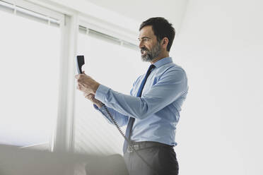 Angry mature businessman holding telephone receiver in office - MOEF02547