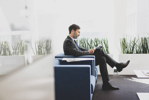 Mature businessman sitting in waiting area reading magazine - MOEF02548