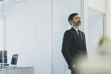 Thoughtful mature businessman leaning against glass wall in office - MOEF02556