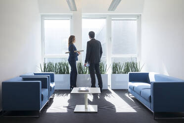 Businesswoman and businessman looking out of window in office lounge - MOEF02569