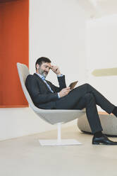 Reifer Geschäftsmann sitzt auf einem Stuhl und benutzt ein Tablet - MOEF02584