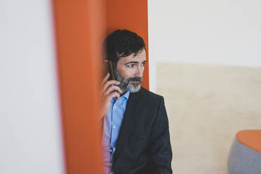Reifer Geschäftsmann sitzt in einer Nische und spricht mit seinem Handy - MOEF02598