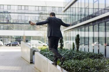 Rückansicht eines Geschäftsmannes, der auf einer Mauer vor einem Bürogebäude balanciert - MOEF02610