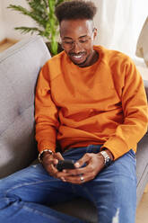 Junger lächelnder Mann sitzt auf einer Couch und benutzt ein Smartphone - IGGF01433