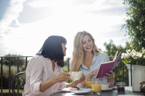 Zwei Frauen frühstücken am Tisch eines Cafés im Freien und lesen ein Buch - AJOF00037