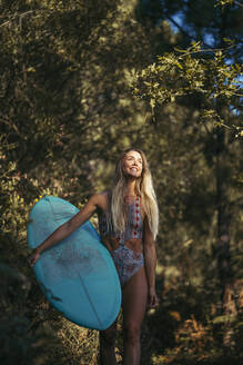 Young blond woman with a surfboard walking in the forest, looking up - MTBF00090