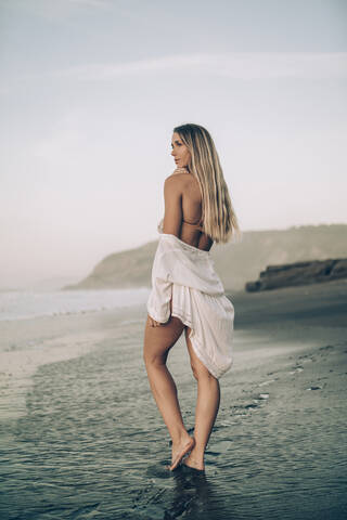 Junge blonde Frau trägt Bikini und weißes Kleid am Strand am Morgen, lizenzfreies Stockfoto