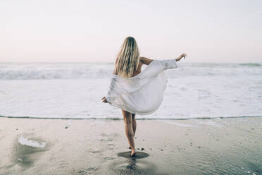 Junge blonde Frau trägt Bikini und weißes Kleid am Strand am Morgen - MTBF00117