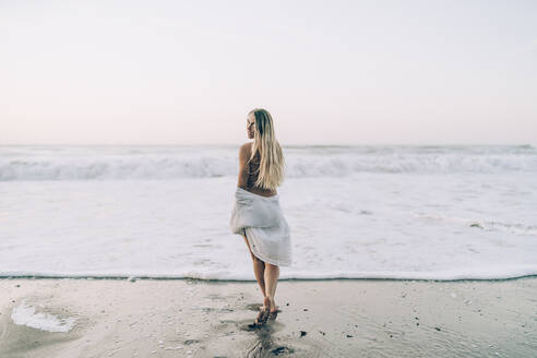 Junge blonde Frau trägt Bikini und weißes Kleid am Strand am Morgen - MTBF00118