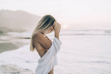 Junge blonde Frau trägt Bikini und weißes Kleid am Strand bei Sonnenaufgang - MTBF00119