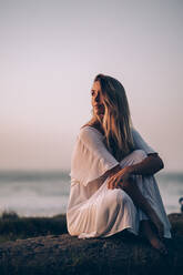 Young blond woman sitting at the beach and looking sideways during sunrise - MTBF00129
