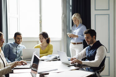 Colleagues disussing project, while mature woman is using smartphone in background - SODF00249