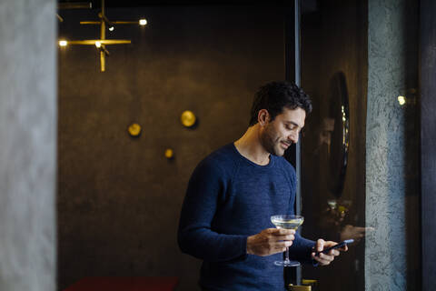 Geschäftsmann in einem stilvollen Büro, der nach der Arbeit einen Cocktail zu sich nimmt und sein Smartphone benutzt, lizenzfreies Stockfoto