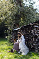 Pregnant woman with dog relaxing at stack of wood in garden - SODF00313