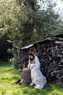 Schwangere Frau mit Hund entspannt sich an einem Holzstapel im Garten - SODF00313