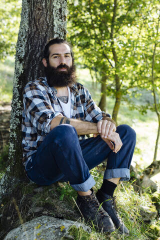 Porträt eines Mannes mit Bart, der an einem Baumstamm im Wald sitzt, lizenzfreies Stockfoto