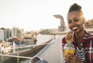Portrait of happy young woman having a drink on rooftop at sunset - UUF19454