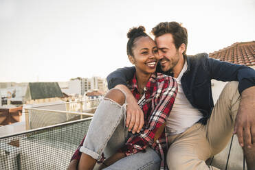 Happy affectionate young couple sitting on rooftop in the evening - UUF19458