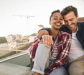 Happy affectionate young couple sitting on rooftop in the evening - UUF19463