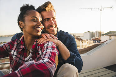 Happy affectionate young couple sitting on rooftop in the evening - UUF19470