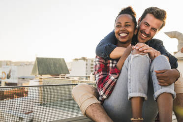 Happy affectionate young couple sitting on rooftop in the evening - UUF19474