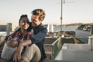 Happy affectionate young couple sitting on rooftop in the evening - UUF19477