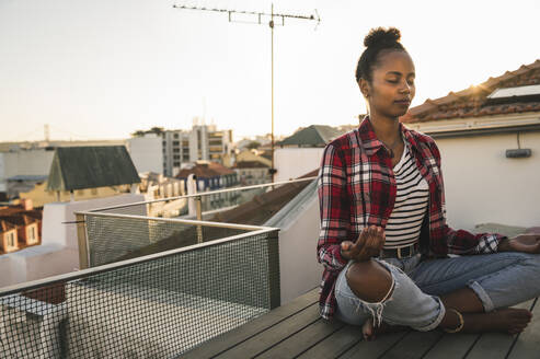 Young woman practicing yoga on rooftop at sunset - UUF19480