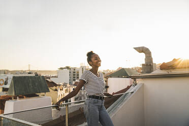 Happy young woman standing on rooftop at sunset - UUF19482