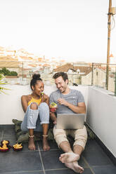 Happy young couple with laptop sitting on rooftop in the evening having a snack - UUF19485