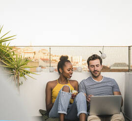 Young couple with laptop and earphones sitting on rooftop in the evening - UUF19487