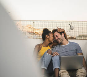 Happy young couple with laptop and earphones sitting on rooftop in the evening - UUF19489