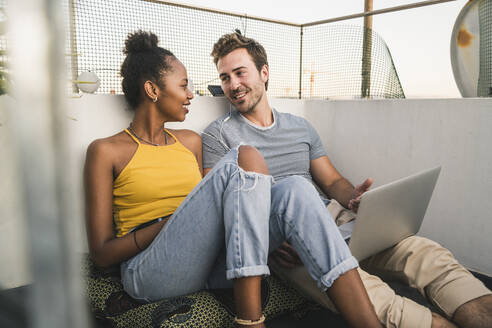 Young couple with laptop and earphones sitting on rooftop in the evening - UUF19490