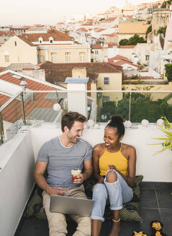 Glückliches junges Paar mit Laptop und Kopfhörern, das abends auf einem Dach sitzt, Lissabon, Portugal, lizenzfreies Stockfoto