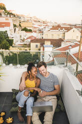 Happy young couple with laptop sitting on rooftop in the evening, Lisbon, Portugal - UUF19493