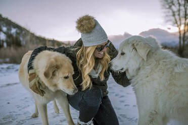 Glückliche Frau, die Hunde streichelt, während sie auf einem verschneiten Feld gegen den Himmel bei Sonnenuntergang kauert - CAVF68281