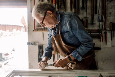 Ein älterer Mann kratzt einen Fensterrahmen auf einer Werkbank in der Werkstatt eines Geschäfts - MASF14019