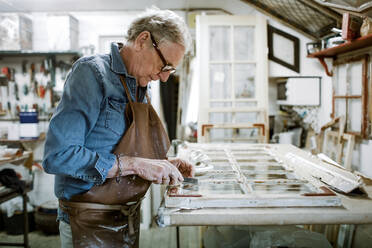 Side view of senior male owner scraping window frame on workbench at store workshop - MASF14027