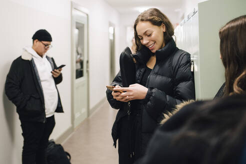 Lächelnder Teenager-Schüler, der sein Smartphone im Korridor benutzt - MASF14071