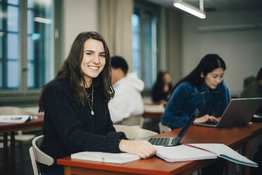 Porträt eines lächelnden Teenagers, der am Schreibtisch sitzt, mit Klassenkameraden im Hintergrund - MASF14083