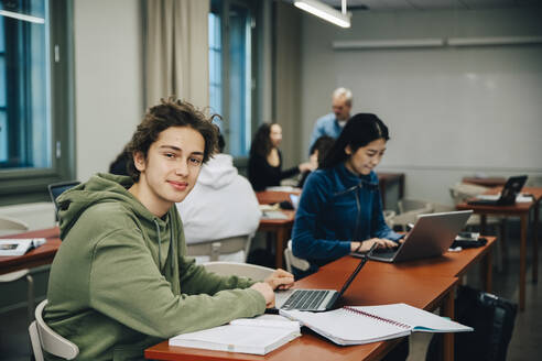 Porträt eines lächelnden Teenagers, der einen Laptop am Schreibtisch benutzt und mit Klassenkameraden im Klassenzimmer lernt - MASF14085