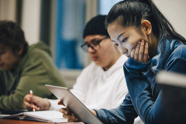 Teenager-Mädchen mit Hand am Kinn, das ein digitales Tablet benutzt, während es neben Klassenkameraden in der High School sitzt - MASF14093