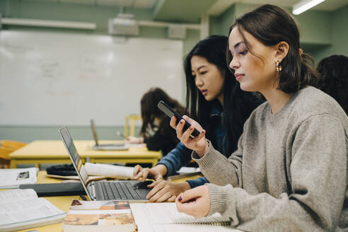 Eine Gymnasiastin benutzt ein Smartphone, während sie neben einem Klassenkameraden sitzt und am Laptop im Klassenzimmer lernt - MASF14109