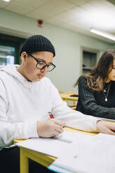 Teenage boy writing on book while studying at desk in classroom - MASF14110