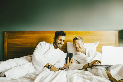 Lächelnder Mann zeigt seinem Freund, der einen Laptop benutzt, sein Smartphone, während er sich im Hotel ins Bett legt, lizenzfreies Stockfoto