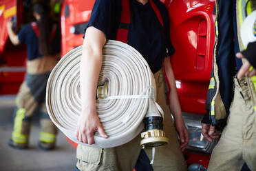 Midsection of female firefighter holding rolled up fire hose while standing with coworker at fire station - MASF14170