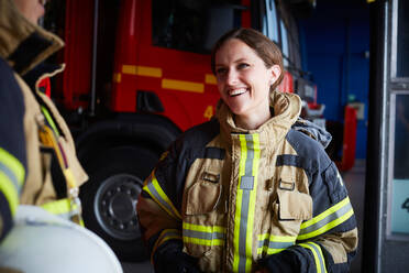 Smiling female firefighter looking at coworker while communicating in fire station - MASF14193