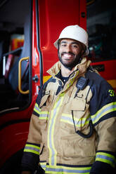 Portrait of smiling male firefighter standing against fire engine at fire station - MASF14201