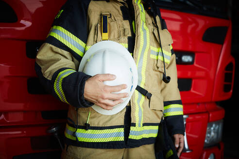 Mittelteil eines weiblichen Feuerwehrmanns, der einen Helm hält, während er auf der Feuerwache steht - MASF14209